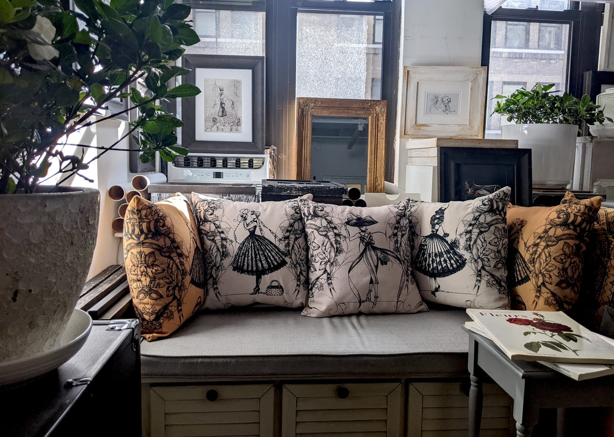 A set of decorative throw pillows arranged on a bench in a styled room. The pillows include a light beige and white patterned design with black dress illustrations of MeowCleeva alongside two mustard yellow pillows with black vintage floral prints.