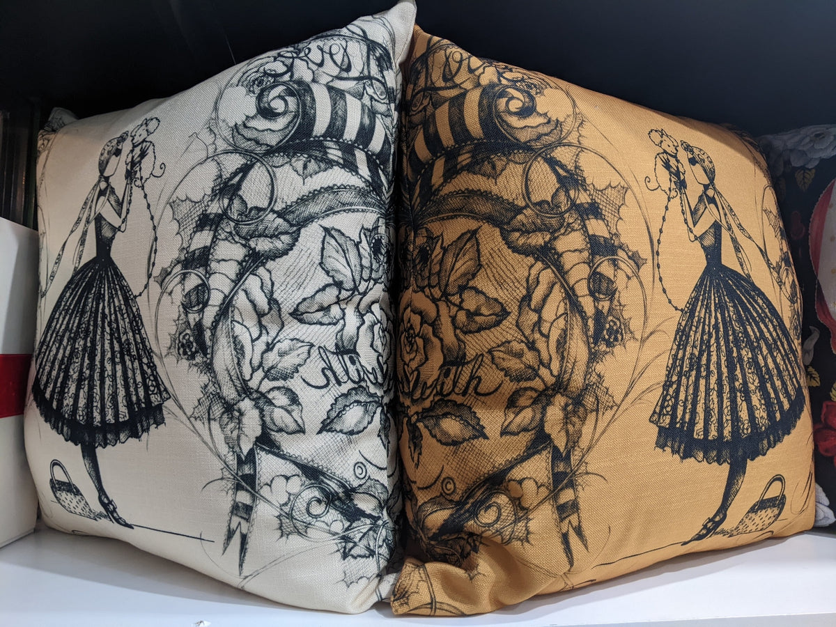 Two decorative throw pillows displayed side by side. The left pillow is cream with a black vintage-style illustration of  the character MeowCleeva® in a skirt and floral scrollwork.