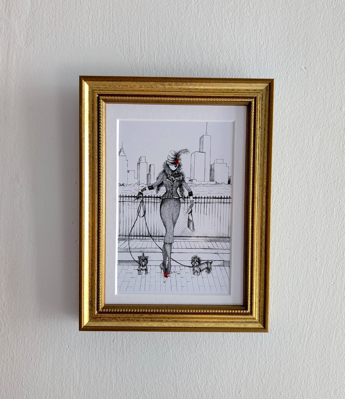 A framed art print featuring a black and white sketch of a woman with a red hat and red shoes, enclosed in a gold frame and hung on a white textured wall.
