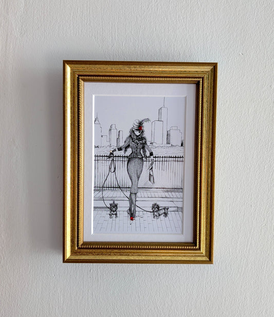 A framed art print featuring a black and white sketch of a woman with a red hat and red shoes, enclosed in a gold frame and hung on a white textured wall.