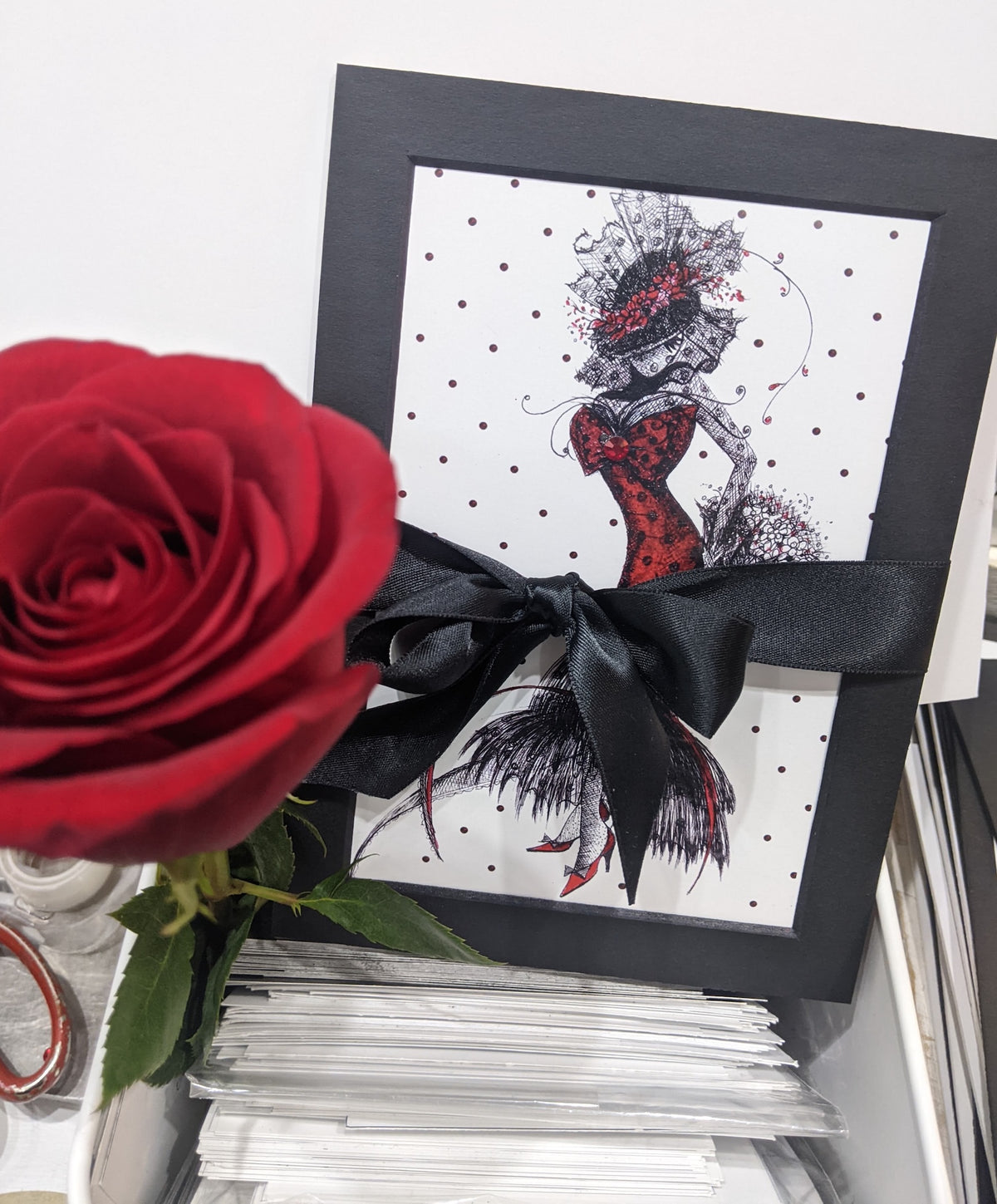 Framed artwork of meowcleeva in a red dress with a black bow, next to a red rose set within prints in a workroom setting 