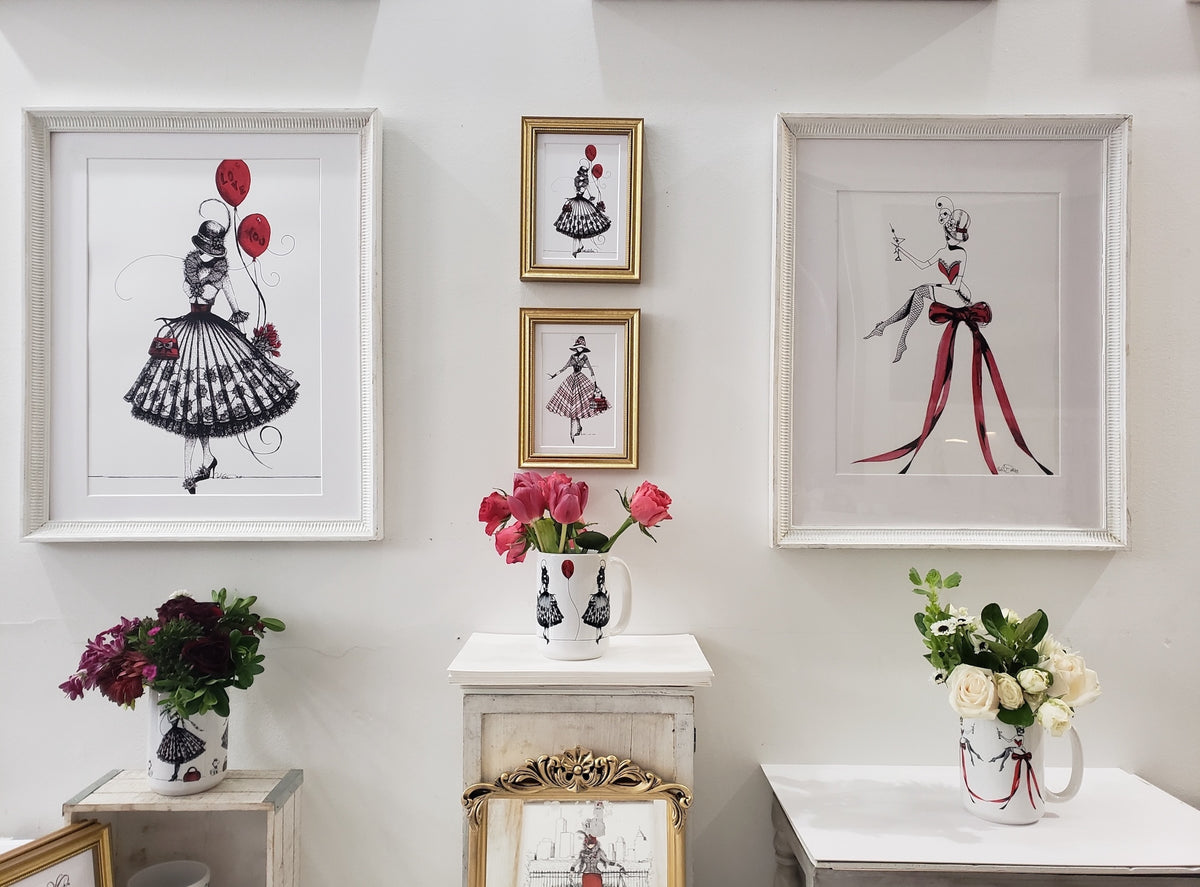 Four framed fashion illustration prints featuring stylized MeowCleeva in black and red dresses. Gallery wall above decorative tables with flowers.