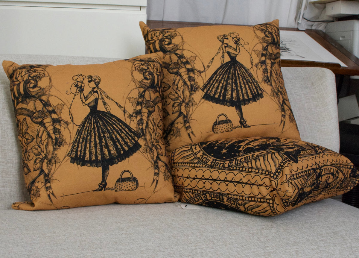 Three decorative throw pillows with a vintage black fashion illustration print of the character MeowCleeva® on a warm mustard-yellow background, arranged on a light gray fabric sofa